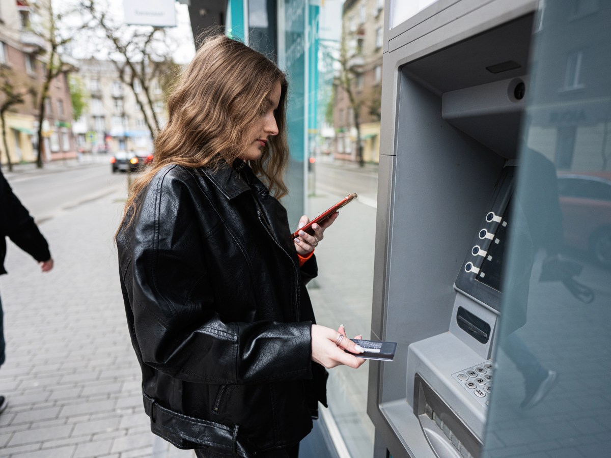 Young woman in leather jacket inserting a credit card to ATM outdoor, while looking on smartphone.