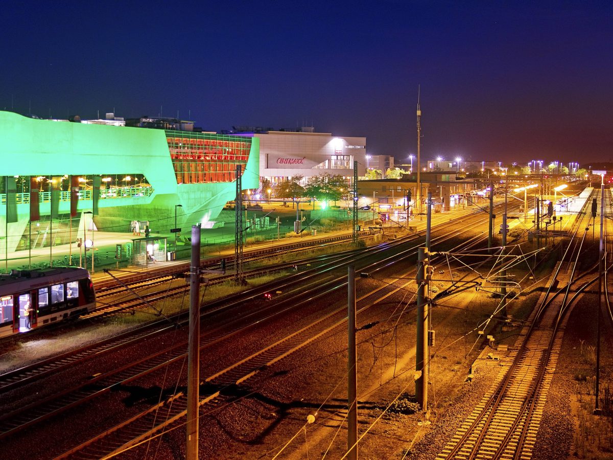 Das bunt illuminierte phæno Gebäude und der Hauptbahnhof mit einem Nahverkehrszug nachts
