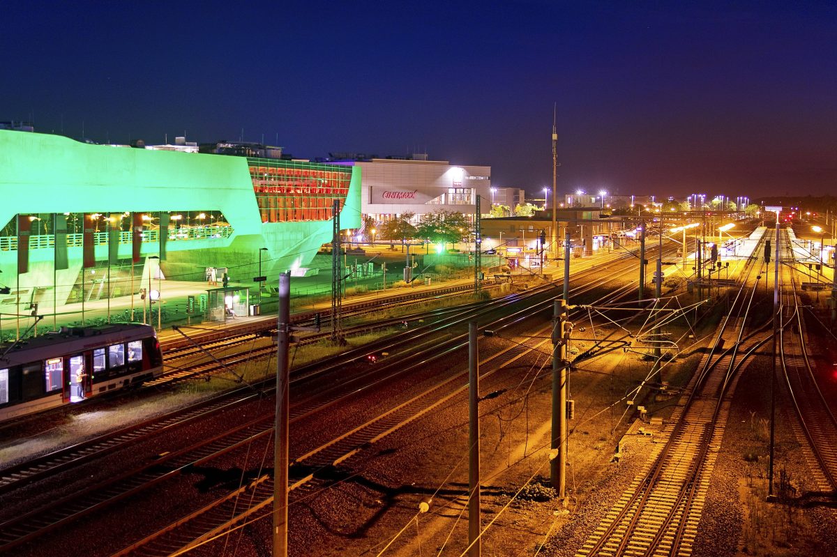 Das bunt illuminierte phæno Gebäude und der Hauptbahnhof mit einem Nahverkehrszug nachts